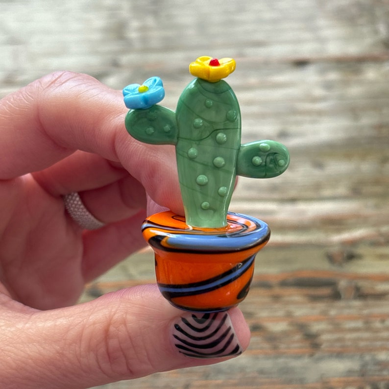 May include: A small glass cactus figurine with green and white details, a blue flower, and a yellow flower with a red center. The cactus is potted in a swirl-patterned orange pot with blue and black accents.