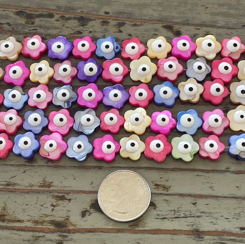 May include: A collection of colorful flower-shaped beads with an evil eye design. The beads are made of mother of pearl and have a white eye with a black pupil. The beads are arranged in rows on a wooden surface.