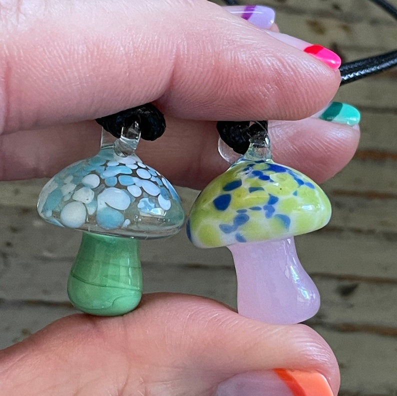 May include: Two glass mushroom pendants on black cords. The mushroom on the left has a blue and white speckled cap and a green stem. The mushroom on the right has a green and blue speckled cap and a pink stem.