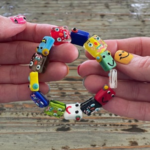 May include: A bracelet composed of multi-colored glass beads, each featuring a unique hand-painted character face. The beads are rectangular and square in shape, showcasing vibrant hues including red, blue, yellow, green, black, and white, strung together.