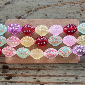 May include: A collection of colorful, seashell-shaped beads in various colors, including pink, red, yellow, and green. Each bead has a textured surface and is adorned with small, contrasting dots. The beads are arranged on a brown rectangular surface.