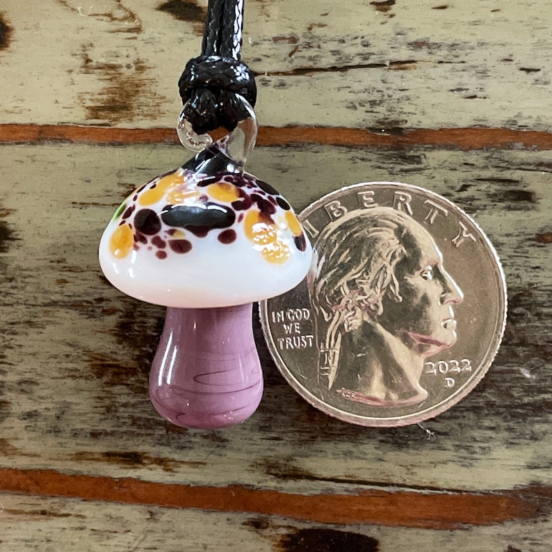 May include: A glass mushroom pendant with a white cap, purple stem, and brown, yellow, and white speckles. The pendant is hanging from a black cord and is next to a US quarter for size comparison.