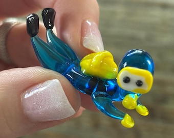 May include: A small glass figurine of a scuba diver wearing a blue wetsuit and yellow scuba gear. The diver is in a mid-air pose, as if they are about to dive into the water.