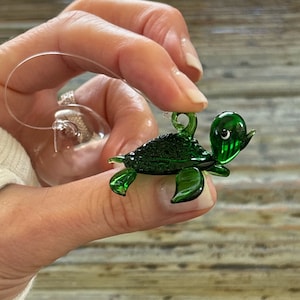 May include: A small, green glass turtle ornament with a clear loop for hanging. The turtle has a detailed shell and is held up by a clear string. A clear glass ball ornament is also visible in the background.