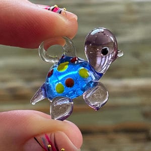 Op de afbeelding: Een miniatuur glazen schildpadfiguur. De schildpad heeft een blauw schild met gele en bruine vlekken. De kop en poten zijn helder en de kop is een doorschijnende paarse kleur. De schildpad wordt tussen twee vingers gehouden.
