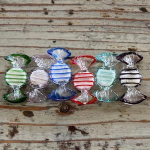 Op de afbeelding: Zeven glazen snoepversieringen in verschillende kleuren, waaronder groen, roze, blauw, rood, turquoise en zwart. Elk ornament heeft de vorm van een ingepakt snoepje met een wit en gekleurd gestreept ontwerp.