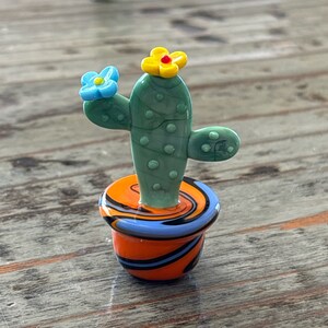 May include: A small green glass cactus with yellow and blue flowers, potted in a swirl pattern orange, blue, and black pot.