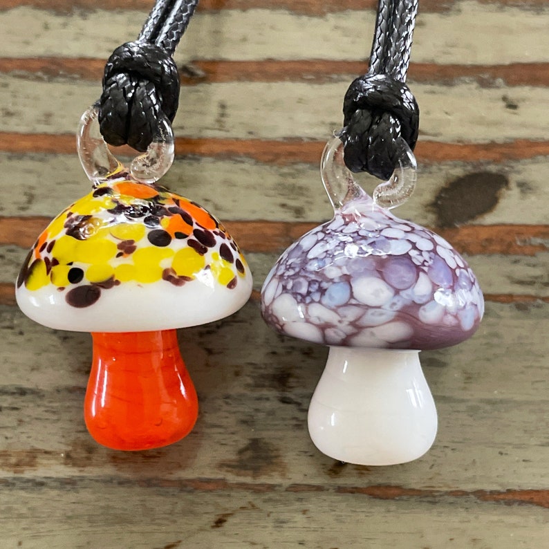 May include: Two glass mushroom necklaces with black cords. One mushroom is white with orange, yellow, and brown spots. The other mushroom is white with purple and blue spots.