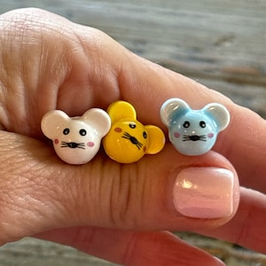 May include: Three small ceramic mouse figurines. The mice are in a hand. The mice are white, yellow, and blue. The mice have black eyes and pink cheeks.