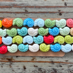 May include: A collection of colorful ceramic seashell beads in various shades of blue, green, red, white, and yellow. The beads are arranged in a row on a brown surface.