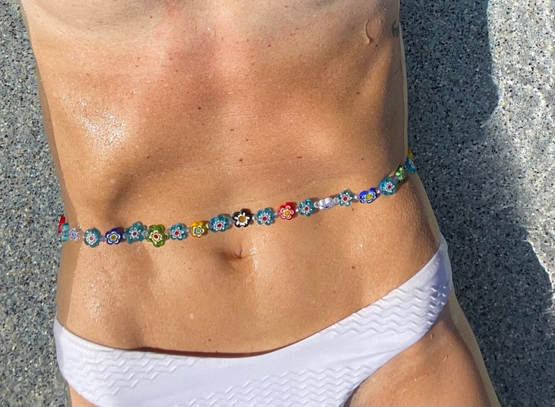 May include: A colorful waist bead strand featuring small, flower-shaped beads in various colors. The beads are strung together, creating a decorative accessory. The image shows the waist beads worn over a white bikini bottom.
