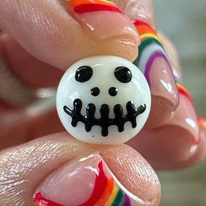 Può includere: Un perlino di vetro bianco con un disegno di teschio nero. Il perlino è tenuto da una mano con smalto per unghie arcobaleno.