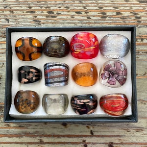 Puede incluir: Surtido de 12 anillos de vidrio con varios patrones y colores, incluyendo rojo, naranja, marrón, negro, plateado y blanco. Los anillos se muestran en una caja negra con un interior blanco.