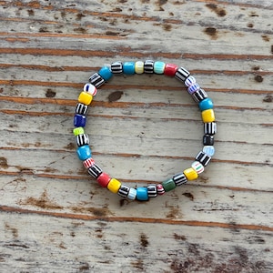 Puede incluir: Una colorida pulsera de cuentas con cuentas cuadradas en tonos azules, amarillos, rojos y rayas blancas y negras. La pulsera es circular y está sobre una superficie de madera.