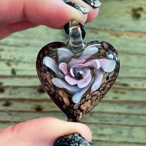 May include: A glass heart pendant with a pink rose design inside. The heart is black with gold flecks and the rose has white and pink petals.