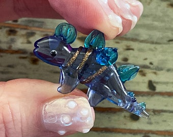 May include: A small glass figurine of a blue and green dinosaur with gold accents. The dinosaur is standing on its hind legs and has a long tail.