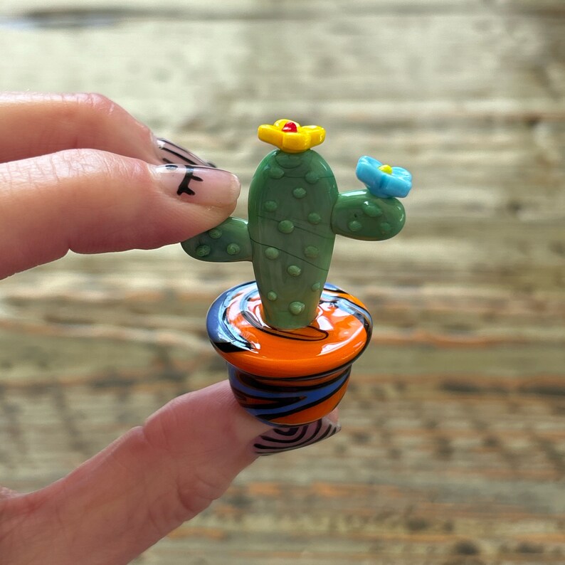 May include: A small glass cactus with a green body, yellow flower, and blue flower, sitting in a colorful orange, black, and blue pot.