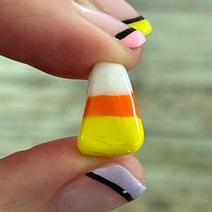 Puede incluir: Un pequeño amuleto de vidrio en forma de maíz dulce con rayas blancas, naranjas y amarillas. El amuleto se sostiene entre dos dedos.