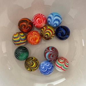 May include: A collection of colorful glass marbles with swirled patterns. The marbles feature a variety of colors, including red, blue, yellow, green, and orange. The marbles are arranged in a white bowl, showcasing their vibrant designs.
