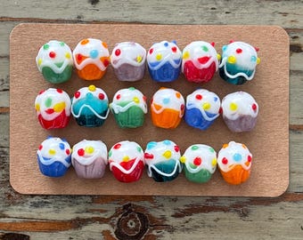 May include: A collection of 16 miniature glass cupcakes with colorful frosting and sprinkles. The cupcakes are arranged in two rows of eight on a brown surface.