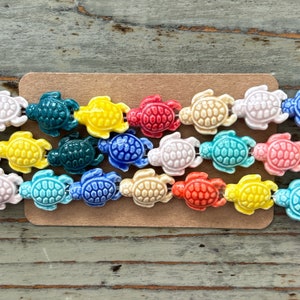 May include: A strand of colourful ceramic turtle beads. The beads are in a rainbow of colours, including yellow, red, blue, green, and pink. The beads are strung on a brown cord and are arranged in a row on a wooden surface.