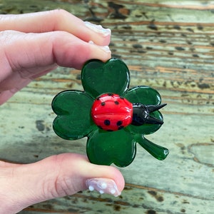 May include: A glass cloverleaf ornament featuring a red ladybird with black spots and a black head. The four-leaf clover is a deep green colour. The ornament is held in a person's hand.
