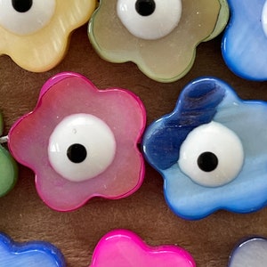 May include: Assortment of colorful flower-shaped beads with an evil eye design. The beads are made of a pearlescent material and come in shades of pink, blue, yellow, and white.