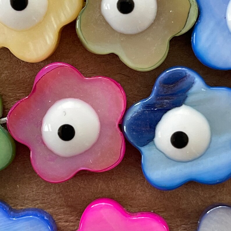 May include: Assortment of colorful flower-shaped beads with an evil eye design. The beads are made of a pearlescent material and come in shades of pink, blue, yellow, and white.