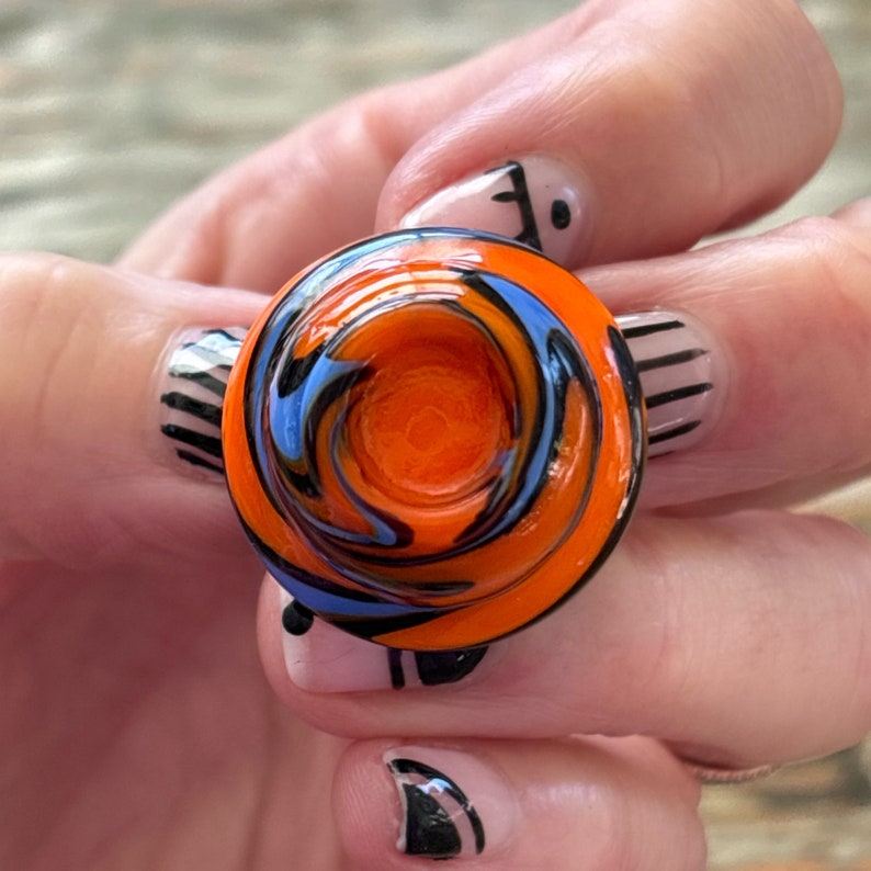 May include: A small, round, orange, blue, and black glass bowl with a swirl pattern. The bowl has a small opening in the center.