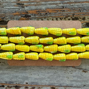 May include: A close-up of a strand of yellow glass beads shaped like corn cobs. The beads have green details to resemble the kernels of corn.