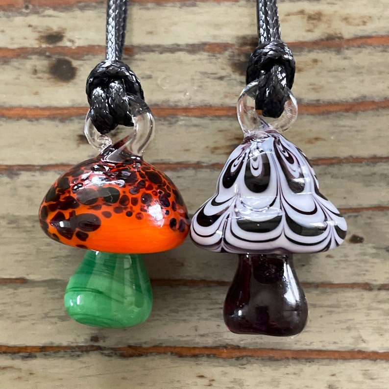 May include: Two glass mushroom pendants on black cords. One mushroom is orange with black spots and a green stem. The other mushroom is white with black swirls and a purple stem.