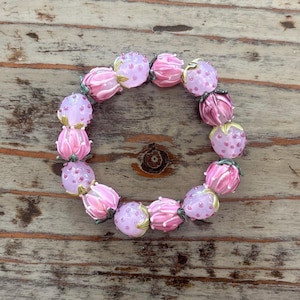 May include: A bracelet featuring pink glass beads shaped like flowers and round beads with pink dots. The bracelet is arranged in a circular shape, showcasing the delicate floral design and color variations. The beads are on a wooden surface.