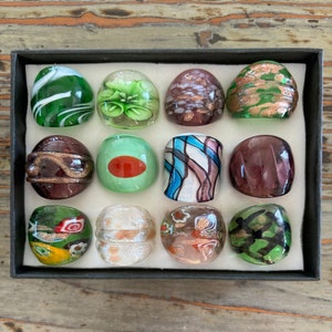 Puede incluir: Un conjunto de 12 anillos de vidrio con varios diseños y colores, incluyendo verde, marrón, blanco y morado. Los anillos están dispuestos en una caja negra con fondo blanco.
