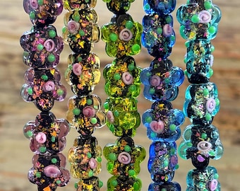 Puede incluir: Primer plano de hilos de cuentas de vidrio de colores. Las cuentas tienen forma de flor con una mezcla de colores que incluyen morado, verde, azul y dorado. Cada cuenta tiene un diseño floral con pequeños acentos rosas y verdes. Las cuentas están ensartadas, creando un patrón vibrante y decorativo.