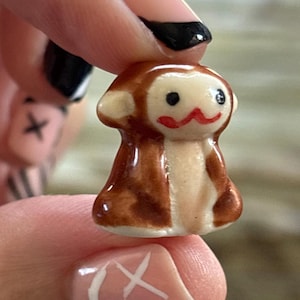 May include: A small, ceramic monkey figurine. The monkey has brown fur, a cream-colored face and belly, and a red mouth. The figurine is held between fingers, showcasing its small size and detailed craftsmanship. The monkey's eyes are black.