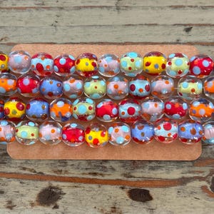 May include: A strand of clear glass beads with colorful polka dots. The beads are oval shaped and have a variety of colors including red, yellow, blue, and green.