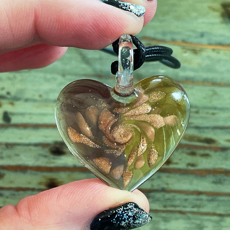 May include: A glass heart pendant necklace with a gold swirl design. The pendant is clear and has a black cord.