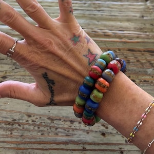 May include: Two beaded bracelets with colorful, round beads in shades of blue, green, red, and orange. The bracelets are stacked on a wrist, with a second bracelet visible. The background is a wooden surface.