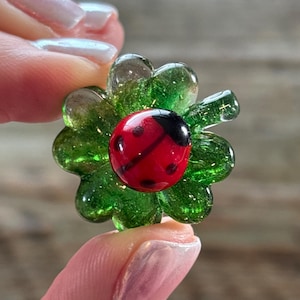 Puede incluir: Una flor de vidrio hecha a mano con un centro de mariquita roja. La flor tiene pétalos verdes con detalles de purpurina. La mariquita es roja con puntos negros y una cabeza negra. La flor se sostiene entre dos dedos.