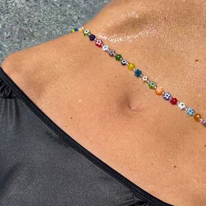 May include: A colorful beaded body chain with flower and eye-shaped beads. The chain is strung across the torso, above a black bikini bottom. The beads are in various colors, including red, yellow, blue, and green.