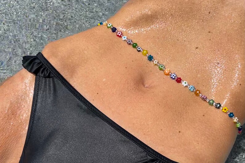 May include: A colorful beaded body chain with flower and eye-shaped beads. The chain is strung across the torso, above a black bikini bottom. The beads are in various colors, including red, yellow, blue, and green.