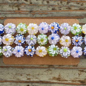 May include: Close-up of a collection of glass flower beads in various colors. The beads are arranged in rows and feature white, blue, yellow, and green petals with yellow centers. The beads are on a brown surface.
