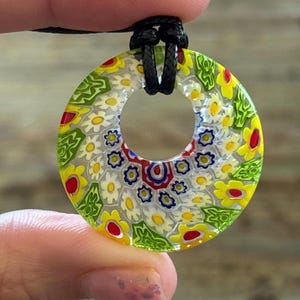 以下が含まれることがあります： 花柄のデザインが施された、カラフルな円形ペンダント。黄色、赤、白、青の花模様が透明な背景に配置されています。黒いコードから吊り下げられています。