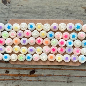 Op de afbeelding: Een verzameling ronde, iriserende witte knopen met een stervormig ontwerp in verschillende kleuren, waaronder roze, blauw, geel en groen.