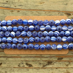 May include: Strands of blue and white swirl-patterned beads. The beads are round and have a glossy finish. The beads are arranged in rows on a brown card, set against a weathered wooden surface. These beads are suitable for jewelry making.