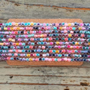 May include: A close-up of a strand of colorful glass beads. The beads are small and round, with a variety of colors and patterns. The beads are arranged in a row on a brown surface.