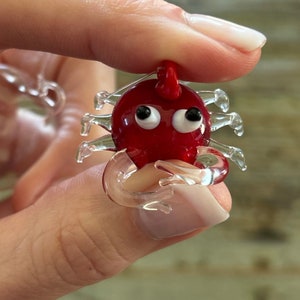 May include: A small, red glass crab figurine with clear glass legs and white eyes. The crab is holding its claws together.