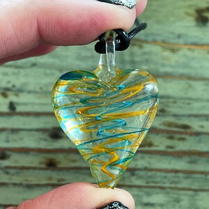 May include: A glass heart pendant with a black cord. The heart is clear with swirls of yellow and blue glass.