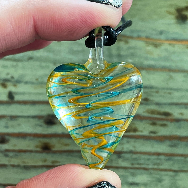 May include: A glass heart pendant with a black cord. The heart is clear with swirls of yellow and blue glass.