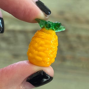 May include: A small glass pineapple charm with a yellow body and green leaves. The charm is about the size of a fingernail.